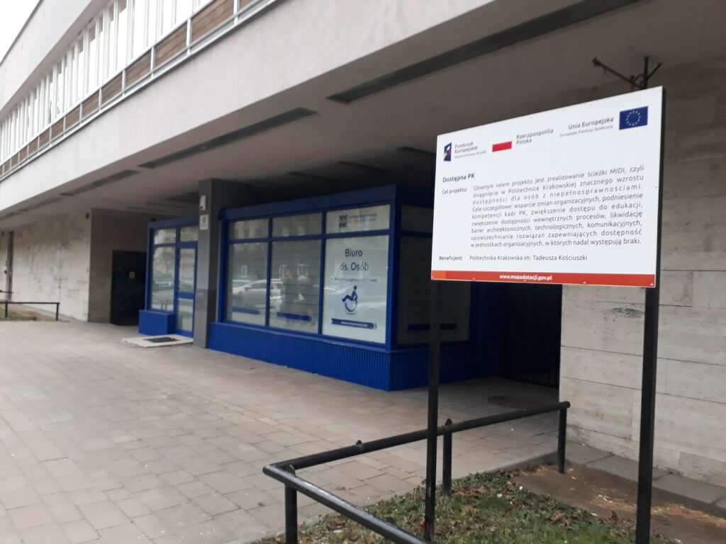 Tablica informacyjna projektu Dostępna PK, w tle niebieski pawilon Biura ds. Osób z Niepełnosprawnościami PK