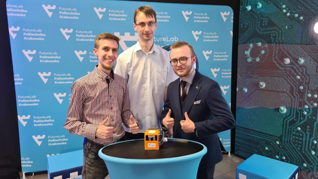 3 studentów - twórców wyświetlacza brajlowskiego pozuje ze stworzonym urządzeniem, w tle baner organizacji FutureLab PK