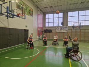 Osoba siedząca na wózku sportowym rzuca piłkę do kosza, 4 zawodników obserwuje